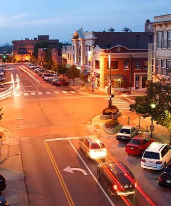 A vibrant downtown street at sunset with a variety of buildings housing shops and businesses. Cars are parked along the sides and streetlights cast a warm glow. The street is busy with moving cars, creating light trails in the dim evening light. - Hausion