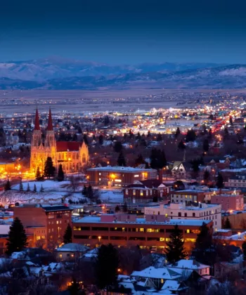 A panoramic view of a city at dusk, with buildings and streets lit up. A prominent illuminated cathedral stands out in the middle of the scene. Snow-covered rooftops and distant mountain ranges are visible under a deep blue sky. - Hausion