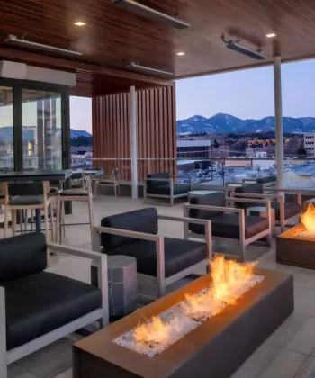 A modern rooftop lounge with contemporary outdoor furniture centered around rectangular fire pits. The space features tall tables and chairs along with cushioned seating. The backdrop includes a view of a cityscape and distant mountains under a dusk sky. - Hausion