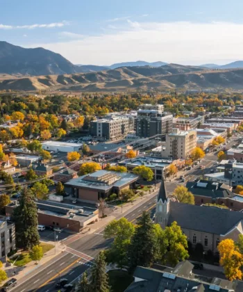 Aerial view of a city nestled in a valley with a backdrop of rolling mountains. The city features a mix of modern and historic buildings, tree-lined streets, and a noticeable church. Autumn colors are visible, with trees showing vibrant yellow and green foliage. - Hausion