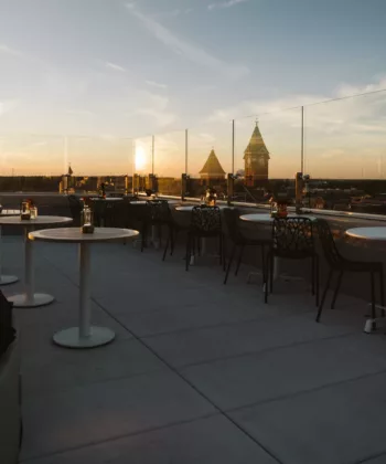 A rooftop terrace at sunset with white tables and dark chairs arranged along the glass barrier. The sky is tinged with orange and blue hues, and there are distant cityscape views with a prominent tower and spire, all contributing to a serene atmosphere. - Hausion