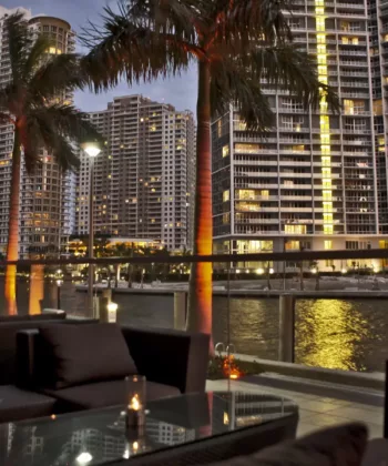 Cozy outdoor seating area by the water with modern glass tables and cushioned chairs, illuminated by small candles. The view includes tall, lit-up skyscrapers and palm trees reflecting in the water at dusk, creating a serene urban atmosphere. - Hausion