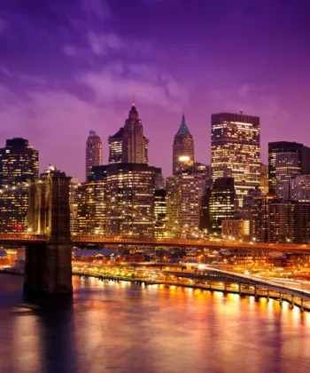 A stunning view of a city skyline at dusk, featuring illuminated skyscrapers and the brightly lit Brooklyn Bridge spanning the river. The sky is a deep purple, casting a serene glow over the scene and reflecting off the water below. - Hausion