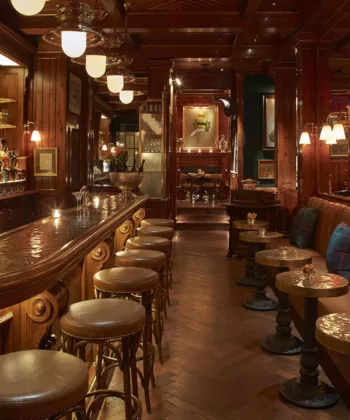 A dimly lit, elegant bar with dark wood paneling and plush leather seating. The curved bar features ornate details with barstools lined up. Tables and couches to the right enhance the cozy atmosphere, illuminated by soft lights hanging from the ceiling. - Hausion