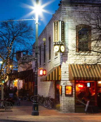 A busy street corner at dusk in a downtown area. A brightly lit restaurant with awnings and signs occupies the corner, displaying "Open" signs. Bikes are parked outside, and string lights are wrapped around leafless trees. Traffic lights and light trails from cars are visible. - Hausion