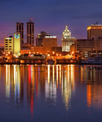 A nighttime cityscape view of Peoria, Illinois, with illuminated buildings reflecting in the Illinois River. The sky has a purple hue, and city lights cast colorful reflections on the calm water. Prominent structures, including the Caterpillar building, are visible. - Hausion