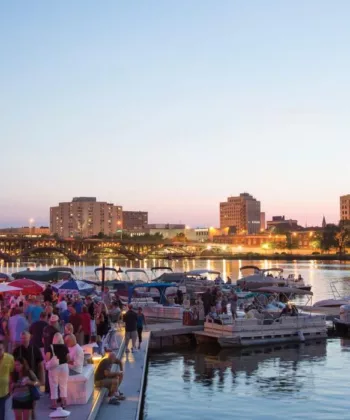 A busy riverside scene at twilight, featuring a crowd of people socializing on a dock and nearby boats. The city skyline is visible in the background, with various buildings and a bridge lit up against the evening sky. - Hausion