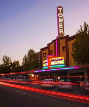 A vibrant street scene at dusk featuring an illuminated theater marquee reading "TOWER" with neon lights. Car light trails streak through the street, and trees line the sidewalk, enhancing the charming, lively atmosphere of the area. - Hausion