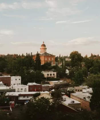 Aerial view of a small town featuring a historic courthouse with a dome at its center, surrounded by trees and old buildings. The weather is clear with a few clouds in the sky, and the scene has a peaceful, rural atmosphere. - Hausion