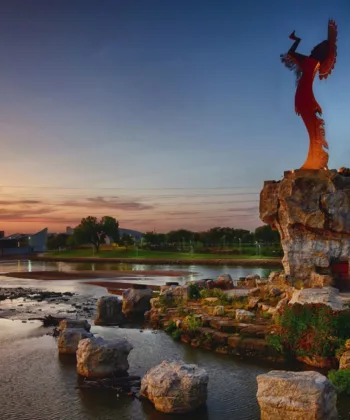 A scenic view at sunset of the Keeper of the Plains sculpture perched on a rocky outcrop above the flowing Arkansas River in Wichita, Kansas. The surrounding area features lush greenery, a walking path, and a vibrant sky transitioning to dusk. - Hausion