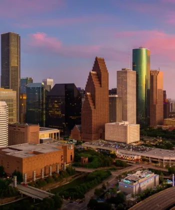 Aerial view of a cityscape during sunset, showcasing modern high-rise buildings with unique architectural designs. The sky is adorned with pink and purple hues. A Ferris wheel and a busy highway are visible in the foreground, adding vibrancy to the urban scene. - Hausion