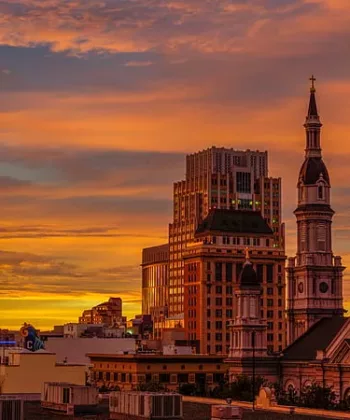 A city skyline at sunset featuring a mix of modern and historic architecture. Prominent buildings include a tall, glass skyscraper with a unique design, a church with a domed roof and a spire, and other varied structures beneath a vibrant, orange sky. - Hausion