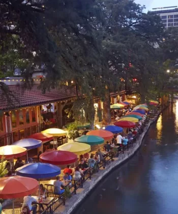 A lively riverside scene at dusk, featuring colorful umbrellas covering outdoor tables along a restaurant-lined walkway. The river reflects the evening lights, and lush trees frame the area. People are dining and strolling along the scenic riverbank. - Hausion