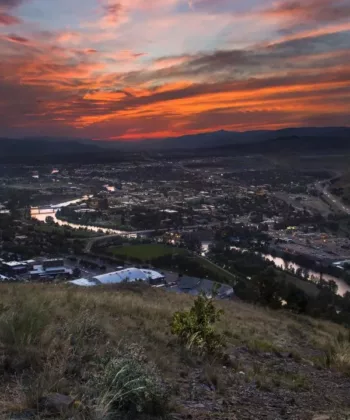 A cityscape at dusk with a vibrant orange and purple sunset. The city is nestled in a valley, surrounded by hills and mountains. A river cuts through the city, reflecting the colors of the sky. Grassy terrain is visible in the foreground. - Hausion