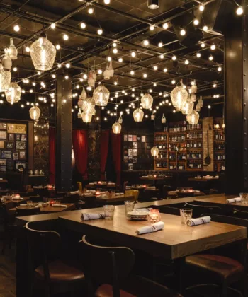 A dimly lit restaurant with warm string lights hanging from the ceiling. The space features wooden tables and chairs, framed photos on the walls, and shelves stocked with bottles. The ambiance is cozy and intimate, with tables set for dining. - Hausion