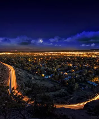 A view of a city at night from an elevated vantage point. Bright city lights illuminate the dense urban area below, with winding roads leading into the city, their paths highlighted by the light trails of moving vehicles. A dark, partly cloudy sky looms above. - Hausion