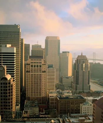 An aerial view of a cityscape at sunrise, featuring tall skyscrapers with varying architectural styles. The sky is partly cloudy, and a large suspension bridge can be seen in the background, spanning across a body of water. - Hausion