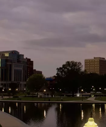 A cityscape at dusk featuring buildings with lights reflecting in a tranquil pond. A paved walkway winds along the water's edge, and a water fountain is visible on the right. The sky is filled with clouds, indicating an approaching evening. - Hausion