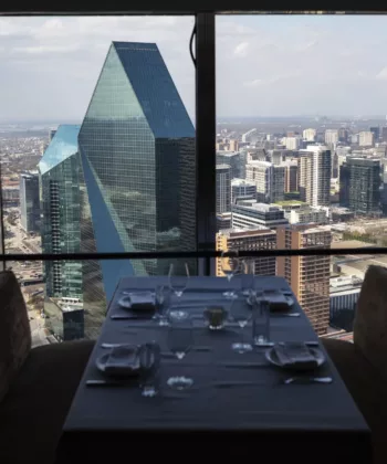 A restaurant table set for four, located by a large window with a panoramic view of a city's skyline. Prominent buildings, including a distinctive green glass pyramid-like structure, are visible. The table has yellow lamp shades and is elegantly arranged. - Hausion