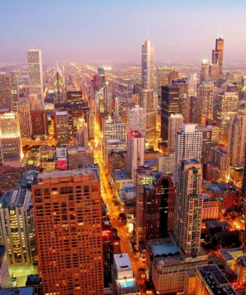 Aerial view of a vibrant city skyline during twilight. Numerous tall buildings, illuminated windows, and streets with traffic lights create a vivid urban landscape. A large body of water is visible to the left, and the sky is a blend of pink and blue hues. - Hausion