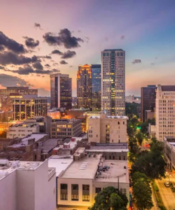 A cityscape at sunset featuring a skyline of tall buildings and skyscrapers. The sky is filled with vibrant colors, transitioning from orange near the horizon to deep blue at the top. Lights illuminate the buildings and streets below, creating a lively urban scene. - Hausion