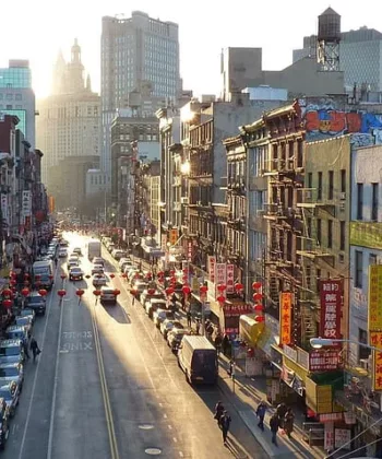 A bustling street in a city during sunset, lined with tall buildings and bustling with traffic and pedestrians. Storefronts with colorful signs and graffiti-covered walls are visible, and the street recedes into the distance, framed by an urban skyline. - Hausion