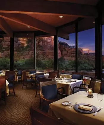 A cozy restaurant with elegantly set tables, each adorned with candles, plates, and glasses. The floor-to-ceiling windows offer stunning views of a rocky desert landscape at dusk, with the sky painted in deep blue and purple hues. - Hausion