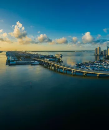 Aerial view of a coastal city at sunset. A bridge spans across the water connecting parts of the city. Skyscrapers and buildings line the waterfront on the right, while a ship is docked near a harbor in the center. The sky is blue with scattered clouds. - Hausion