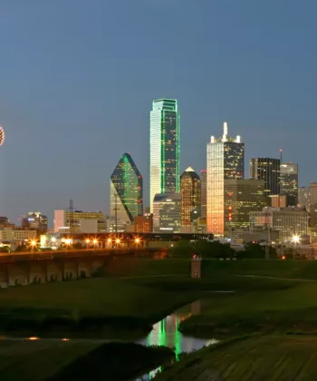 A nighttime view of the Dallas skyline, showing illuminated skyscrapers including the green-lit Bank of America Plaza and Reunion Tower with its spherical lights. The city lights reflect on the river and the sky subtly transitions from dusk to night. - Hausion