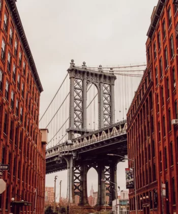 A view of the Manhattan Bridge framed by two rows of red-brick buildings on either side. The iconic bridge spans the background while the brownstone buildings, featuring fire escapes, line the street, creating a classic urban scene under a cloudy sky. - Hausion