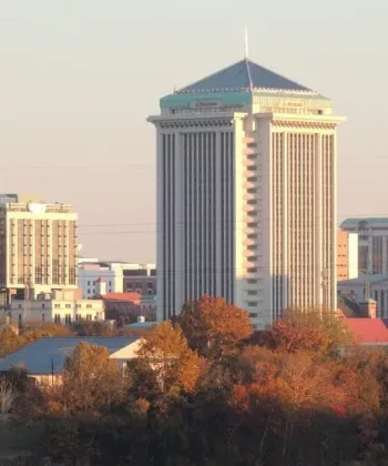 A cityscape featuring a prominent, tall, cylindrical building with a pointed roof at the center, surrounded by other modern buildings. In the foreground, there are trees with autumn-colored foliage, and everything is bathed in a soft, warm light. - Hausion