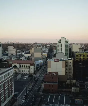 An aerial view of a cityscape at dusk. The scene shows a mix of modern and older buildings, including high-rises and shorter structures. The streets are lined with vehicles and trees, with a slightly pinkish hue in the sky. - Hausion