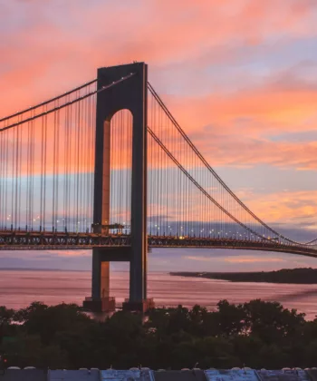 A suspension bridge stretches across a body of water during sunset. The sky is painted with shades of orange, pink, and purple. Trees and buildings are visible in the foreground, while the horizon line meets the glowing sky. - Hausion