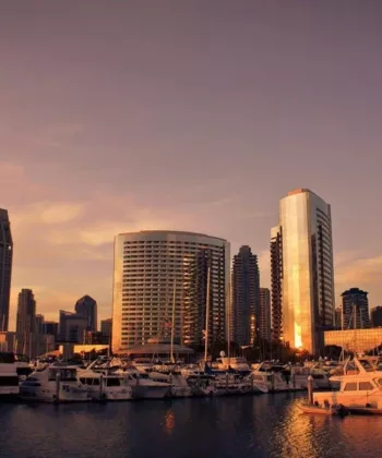 Sunset view of a city marina with several docked boats. The scene includes tall, modern buildings reflecting the golden light of the setting sun, with a clear sky and a peaceful body of water in the foreground. - Hausion