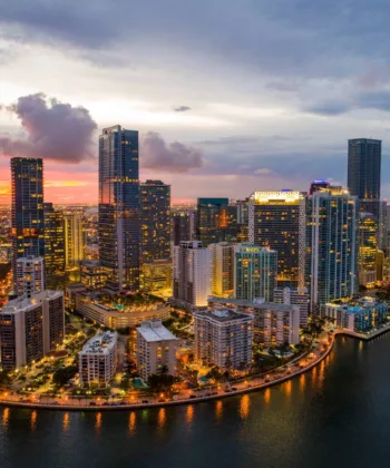 Aerial view of a vibrant cityscape at dusk, showcasing illuminated high-rise buildings reflecting in the water. The sky features a mix of blue, orange, and pink hues as the sun sets, casting a warm glow over the bustling urban area. - Hausion