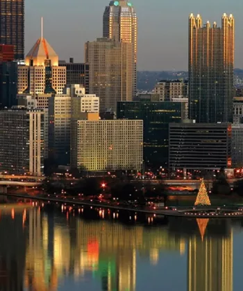 A vibrant cityscape at dusk featuring a skyline with various tall buildings, some illuminated. A river in the foreground reflects the city lights, and a brightly lit triangular structure stands near the water. Bridges and streets are visible amid the city lights. - Hausion