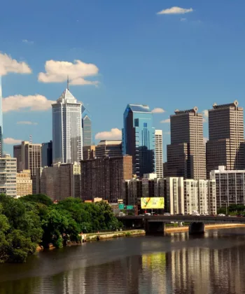 Philadelphia city skyline viewed from across the Schuylkill River, featuring a mix of modern and classic skyscrapers under a clear blue sky. A bridge spans the river, while lush green trees border the water, reflecting the cityscape. - Hausion