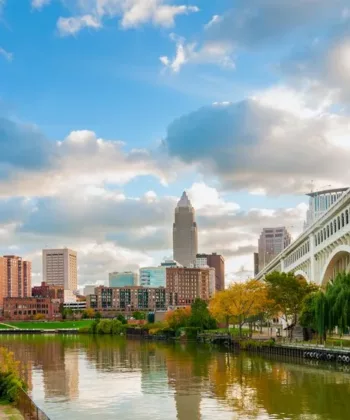 A cityscape view featuring a mix of modern and historic buildings along a river, with a large arched bridge on the right side. The sky is partly cloudy, and trees with autumn foliage line the riverbank. The water reflects the buildings and the sky. - Hausion