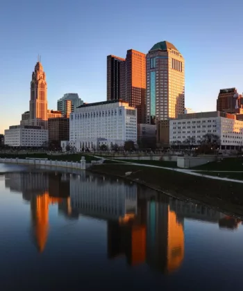 A city skyline at sunset is reflected in a calm river. Tall buildings with a mix of modern and historic architecture are illuminated by the golden light. Green spaces and walkways line the riverbank, enhancing the urban scenery. - Hausion