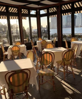 A sunlit café with floor-to-ceiling windows offering views of a river and city skyline. The seating area features small tables covered with white tablecloths and wicker chairs. The warm sunlight creates a cozy and inviting atmosphere inside. - Hausion