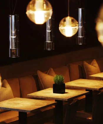 A dimly lit, modern cafe features a row of wooden tables with black chairs and beige cushions against a dark wall. Bright spherical pendant lights hang from the ceiling, casting a warm glow over the space. Small plants in black pots adorn each table. - Hausion