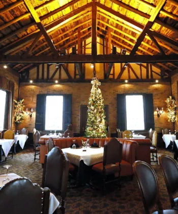 A cozy, rustic restaurant interior with brick walls and a high wooden-beam ceiling. The room is decorated for the holiday season with a large, beautifully adorned Christmas tree at the center. Tables are set with white tablecloths and elegant place settings. - Hausion