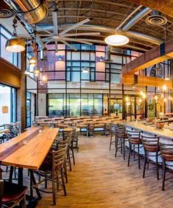 A well-lit, modern restaurant interior featuring wooden tables and chairs. The area includes a long bar on the right with high stools, exposed ductwork, and large windows letting in natural light. The decor blends industrial and rustic elements, creating a cozy atmosphere. - Hausion