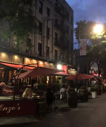 Street scene at dusk with an outdoor dining area in front of a restaurant named "Enzo's." Several people are seated under large red umbrellas. The surroundings include adjacent buildings, trees, and streetlights illuminating the area. - Hausion