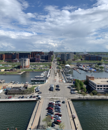 Aerial view of a harbor town showing a wide road stretching into the distance, flanked by buildings and parking spaces. Boats are docked along the water, and the sky is partly cloudy. Green spaces and trees are visible amidst the urban landscape. - Hausion