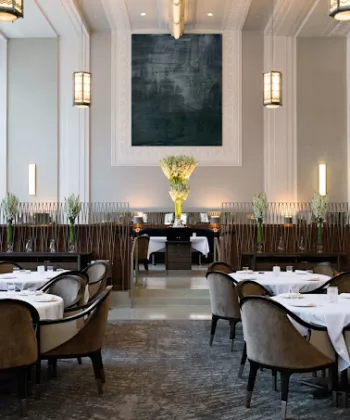 A spacious, elegant restaurant interior with high ceilings, large windows, and modern chandeliers. Round tables with white tablecloths and plush, grey chairs are neatly arranged. A large, dark artwork and numerous flower arrangements add sophistication to the setting. - Hausion