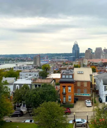 A scenic view of a mid-sized city skyline featuring a cluster of modern buildings. In front, there are older residential homes with green trees dotting the neighborhood. The sky is overcast, and a river can be seen flowing through the city in the background. - Hausion