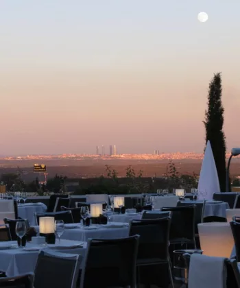 An outdoor restaurant with neatly set tables and chairs overlooks a cityscape under a calming evening sky. A full moon is visible in the sky, casting soft light, and tall buildings can be seen in the distance. Two tall trees frame the view. - Hausion