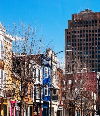 A city street scene with various colorful storefronts and older brick buildings. In the background, a tall modern skyscraper towers over the shorter buildings. Bare trees line the sidewalk under a clear, blue sky. - Hausion