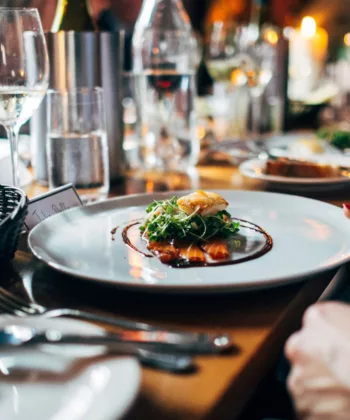 A close-up of a beautifully presented dinner plate featuring a gourmet dish with garnishes on a sauce-drizzled plate at a restaurant. Various wine glasses, a basket of bread, and other dining utensils are seen on the elegantly set table in the background. - Hausion